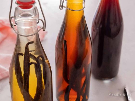 Tiga botol kaca berisi cairan dengan tingkat kepekatan warna berbeda, masing-masing berisi batang vanili.