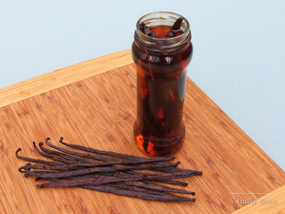 Toples kaca berisi cairan cokelat kemerahan di atas talenan bambu, dengan batang vanili kering di sebelahnya.
