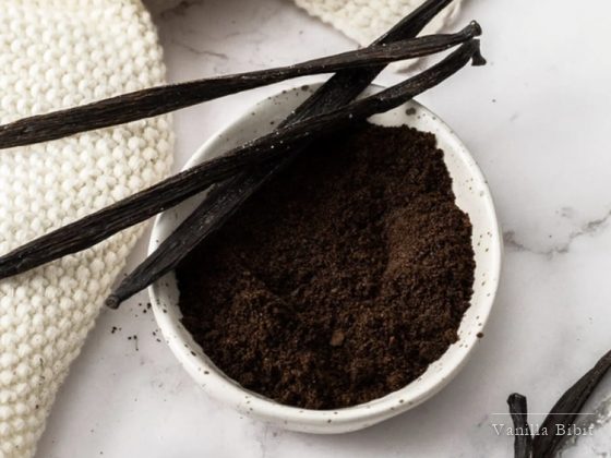 Mangkuk putih berisi bubuk vanili berwarna cokelat tua dengan beberapa batang vanili di sampingnya.